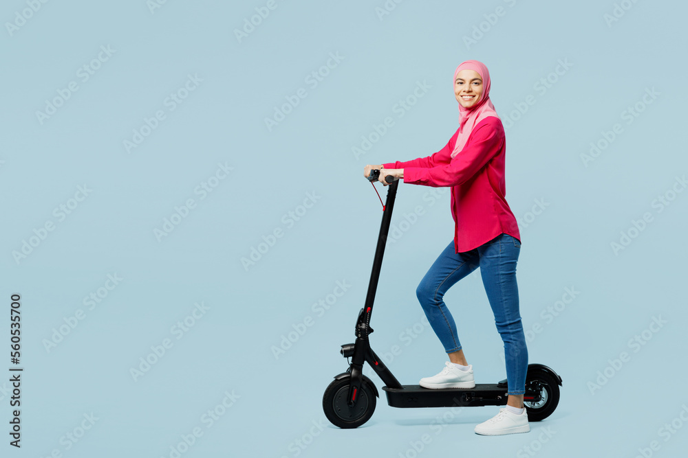 Full body profile young fun arabian muslim woman wear pink abaya hijab riding electric scooter isolated on plain pastel light blue cyan background studio portrait. People uae islam religious concept. Full body profile young fun arabian muslim woman wear pink abaya hijab riding electric scooter isolated on plain pastel light blue cyan background studio portrait. People uae islam religious concept.
