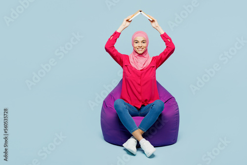 Full body young arabian muslim woman in pink abaya hijab sit in bag chair holb book about head like roof isolated on plain pastel light blue cyan background studio People uae islam religious concept © ViDi Studio
