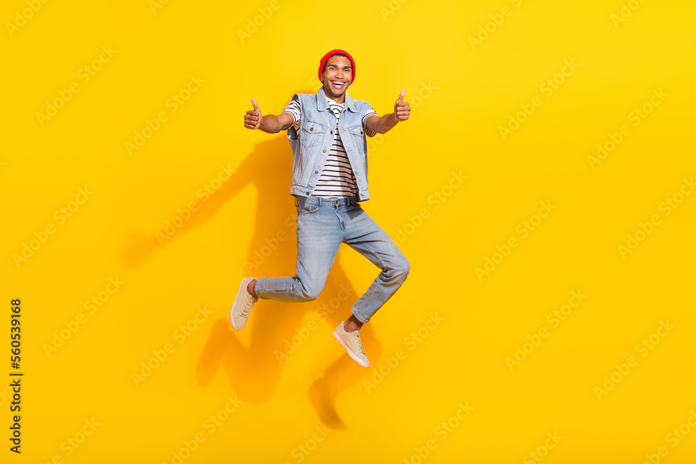 Full length photo of cool cheerful guy dressed denim clothes jumping high showing thumbs up isolated yellow color background