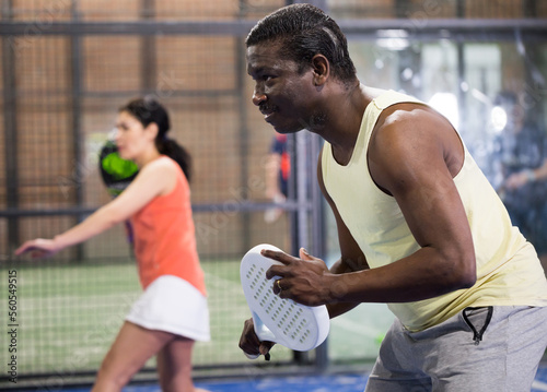 Portrait of sporty adult african american man playing padel on indoor court, ready to hit ball. Sport and active lifestyle concept.