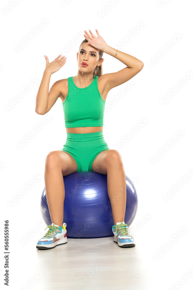 young sporty tired woman in green shorts and top sitting on fitness ball on white background young sporty tired woman in green shorts and top sitting on fitness ball on white background
