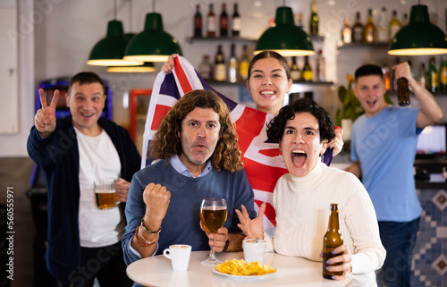 Happy young adult fans waving British flag while drinking beer and watching match together in sports bar