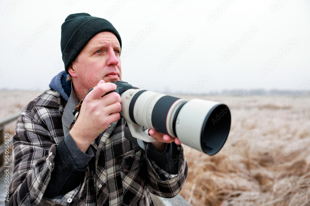 A photographer wearing winter clothing holds a camera and telephoto lens as he looks out across the winter landscape
