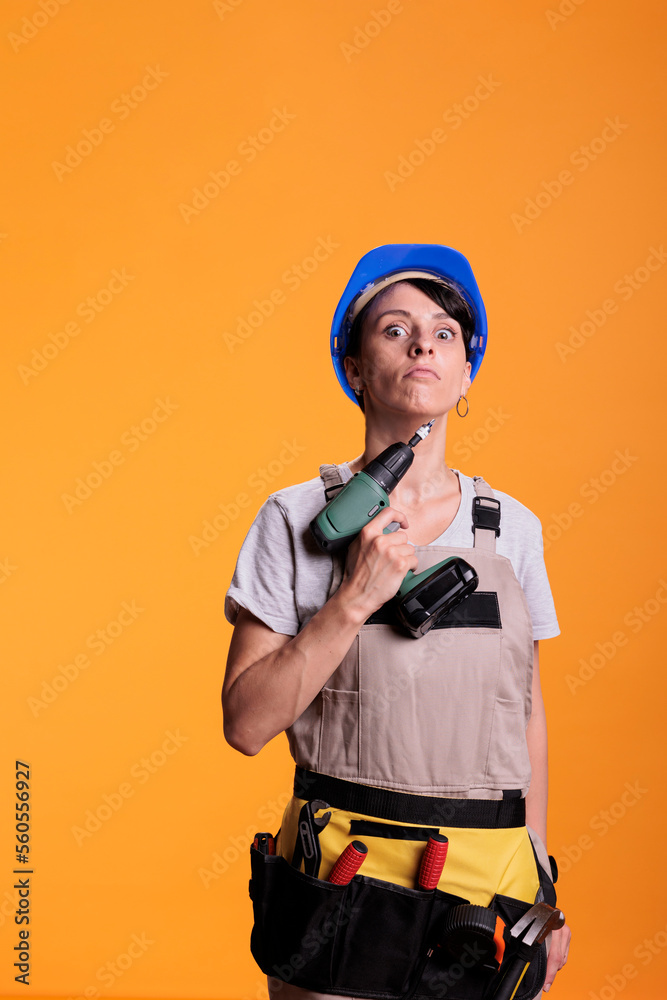 Female repair contractor holding power drill in front of camera, using electric drilling tool before renovation project. Construction worker wearing blue helmet and building uniform. Female repair contractor holding power drill in front of camera, using electric drilling tool before renovation project. Construction worker wearing blue helmet and building uniform.