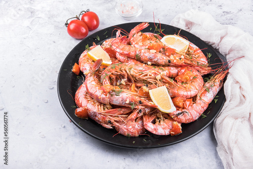 Baked langoustines in garlic sauce with cherry tomatoes on a black plate on a light marble background, side view. 