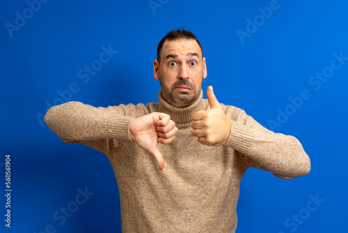 Bearded hispanic man wearing a turtleneck sweater giving a thumbs up and a thumbs down in a gesture of doubt and confusion isolated over blue background.