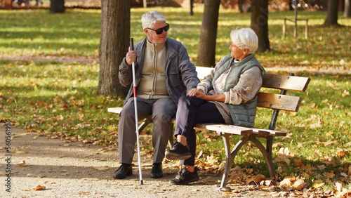 Blind senior man with sunglasses on sitting with his wife on a bench in the park. High quality photo