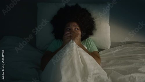 Adult african american woman scared by noise in home or hotel room. Girl with afro hairstyle infantility hiding under blanket, looking around scared to see the monster or ghost, slow motion nightmare photo