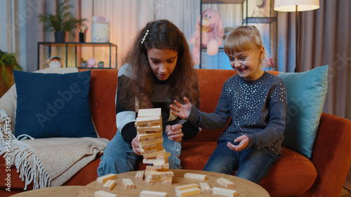 Teenage child and little sister kid girls build tower from wooden bricks, losing board game competition. Siblings children or best friends having fun with tower on table, playing with blocks at home