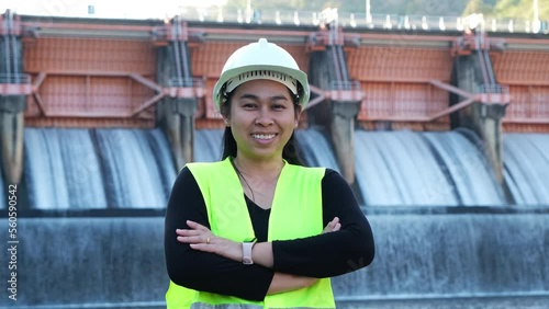 Smiling female engineer in green vest and helmet standing outside against background of hydroelectric dam, floodgate with flowing water through gate.