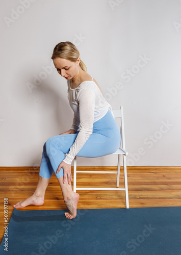 A young woman in blue leggings sits on a chair and holds on to her sore leg (ID: 560597158)