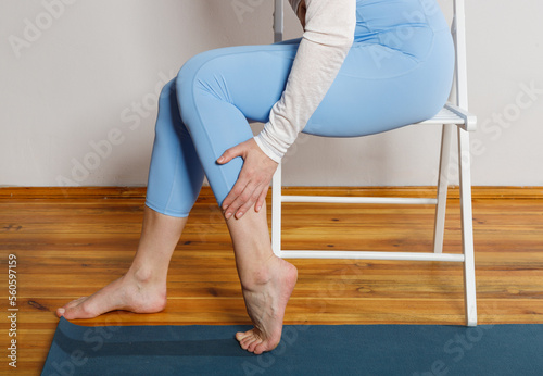 A young woman in blue leggings sits on a chair and holds on to her sore leg (ID: 560597159)