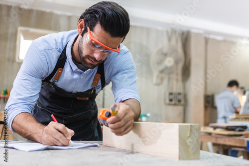 young carpenter caucasian man using measuring tape take note wood size at workspace. craftsman profession in wood factory.