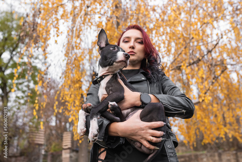 Girl with burgundy hair and in an elegant dress with a cute puppy. Holds a dog in her arms against the background of a tree with yellow leaves. Dog mix: Staffordshire Terrier and Pit Bull Terrier