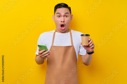 Shocked young Asian man barista barman employee wearing brown apron work in coffee shop holding mobile phoe and paper cup of coffee or tea isolated on yellow background. Small business startup photo