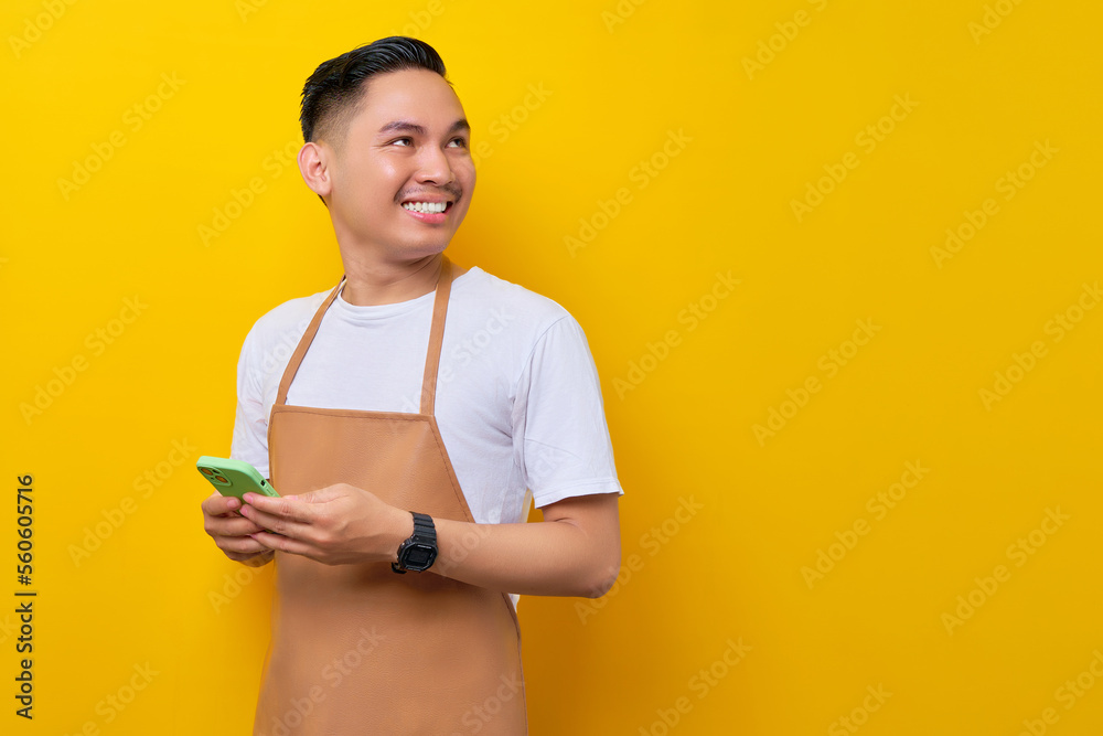 Φωτογραφία smiling young Asian man barista barman employee wearing brown apron work in coffee shop holding mobile phone and looking aside isolated on yellow background