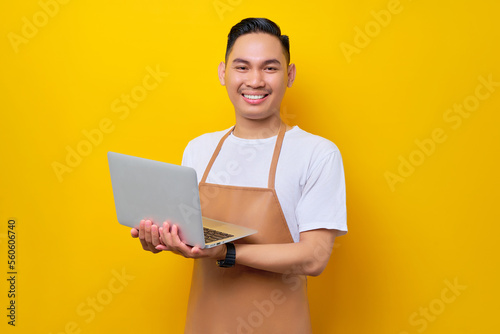smiling young Asian man barista barman employee wearing brown apron work in coffee shop using work on laptop pc computer on yellow background. Small business startup concept