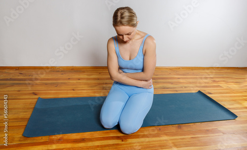 Young woman in a blue solid sports overalls on a yoga mat and holds on to her sore stomach