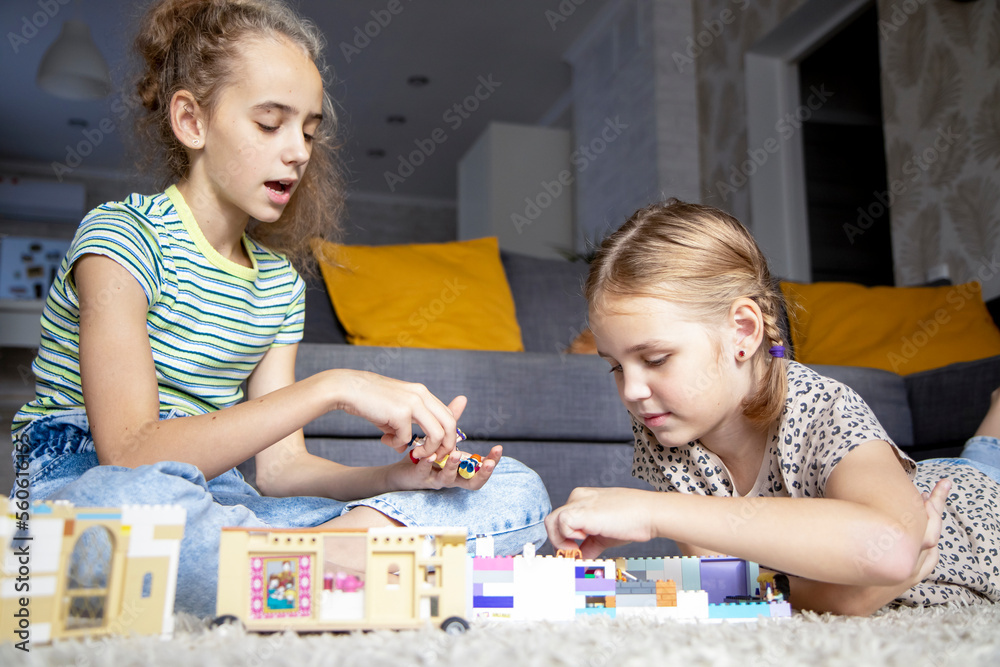 Fototapeta premium Two little sister girls play with multi-colored toy blocks at home in the living room. Educational games for children. Mess in the game room.