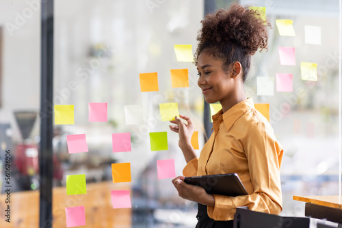 Young Serious african american Creative team use post it notes to share idea sticky note on glass wall. Asian business people design planning and Brainstorming thinking sticky History notes concept. 
