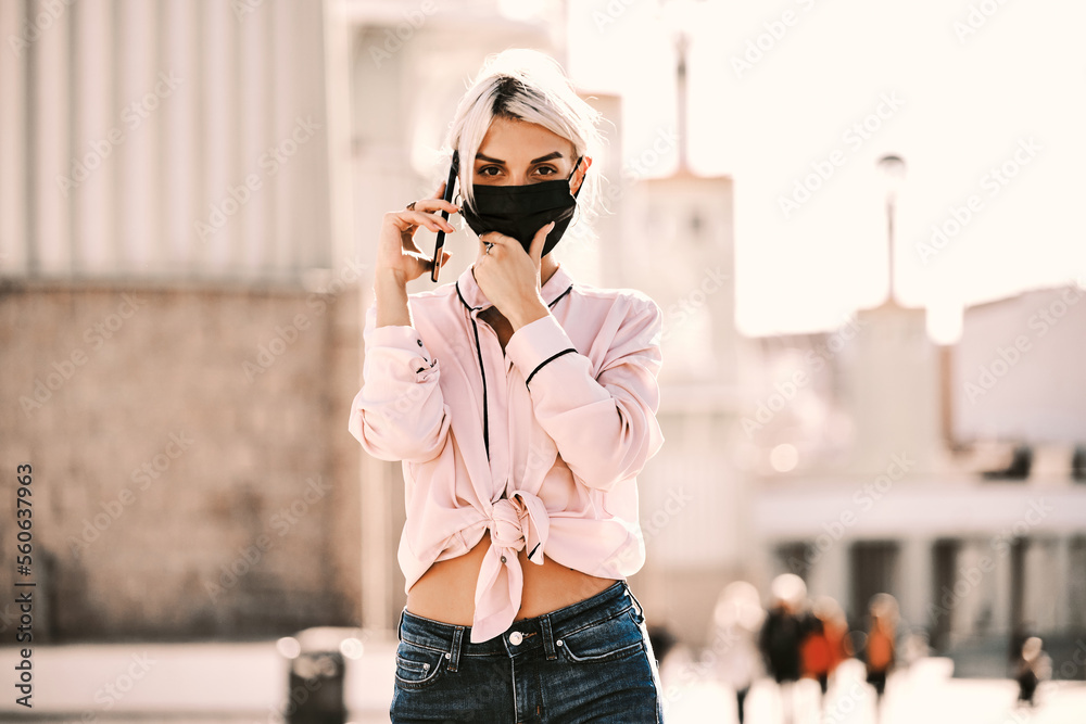 Fototapeta premium Woman with face mask looking at camera while talking on the phone outdoors.