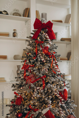  snow covered christmas tree decorated with red decorations indoors. artificial fir tree with yellow garland lanterns and red bows close-up