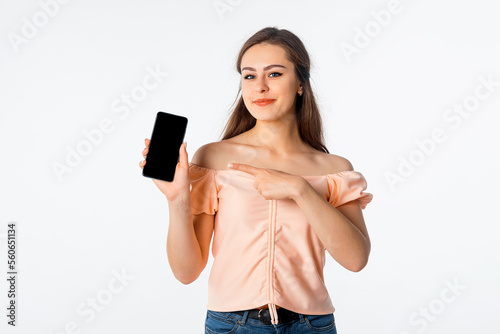 Happy smiling young woman in stylish outfit, pointing at mobile phone screen, showing smartphone display and smiling pleased, white background