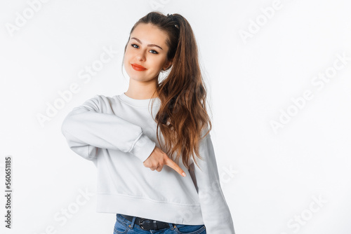 Happy young woman in white sweatshirt, pointing finger down, showing amazing news, promo banner, offer. Smiling brunette female stands against white studio background photo