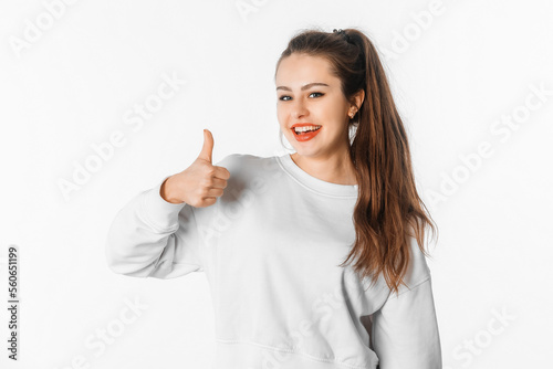 Excited smiling young brunette woman with long hair, wear white sweatshirt show thumb up. Smiling girl looks cheerful and joyful, stands against white studio background
