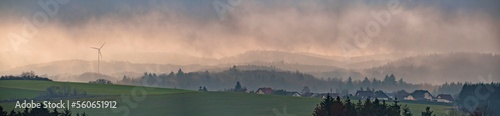 Foggy nature landscape with mountains and wind turbines at village.