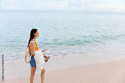 Woman smile with teeth after swimming in the ocean with a backpack in a wet yellow tank top and denim shorts walks along the beach  summer vacation on an island by the ocean in Bali sunset