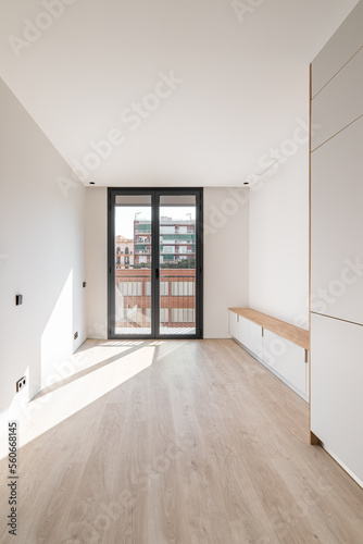 Direct view of black metal framed glass sliding doors at end of an empty bright clean room. Doors leading to spacious balcony overlooking neighboring house. Sunlight enters through glass door.
