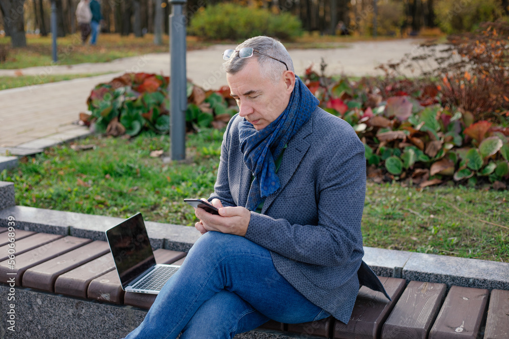 Obraz premium Aged gray haired businessman with glasses sit on bench in autumn city park working on laptop and chatting on telephone.