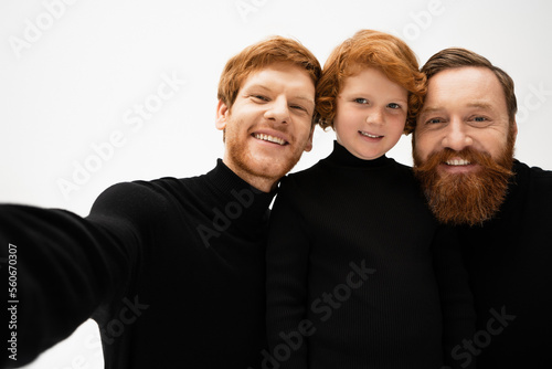 cheerful redhead kid looking at camera near bearded father and granddad in black turtlenecks isolated on grey.