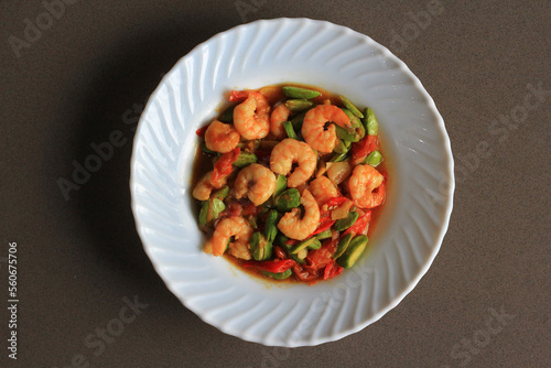 Chili prawns or Sambal Udang with petai or pete or mlanding (Parkia speciosa) cooked with mashed red chili and tomato in white plate, on black background. Indonesia cuisine food, top view.