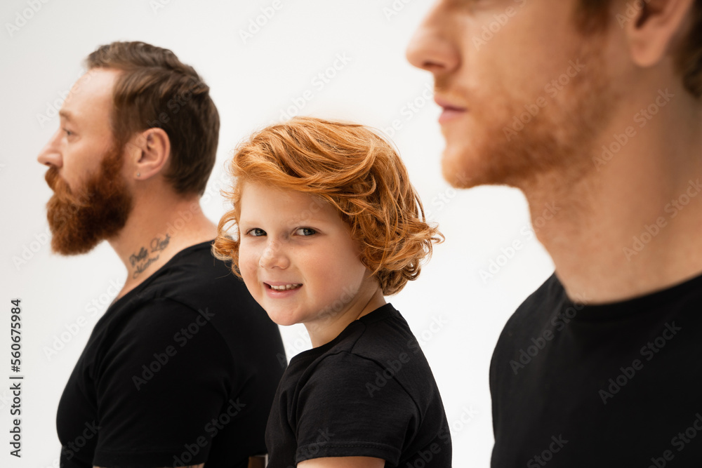 happy red haired boy smiling at camera near bearded tattooed grandpa and dad on blurred foreground isolated on grey. happy red haired boy smiling at camera near bearded tattooed grandpa and dad on blurred foreground isolated on grey.