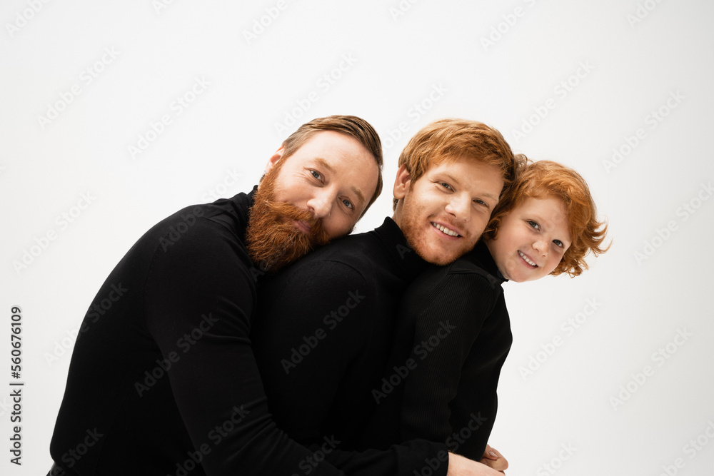 cheerful redhead men and kid in black sweaters leaning on each other and smiling at camera isolated on grey. cheerful redhead men and kid in black sweaters leaning on each other and smiling at camera isolated on grey.