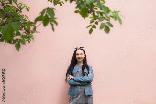 A positive girl with a beautiful smile in a gray dress and a blue denim jacket, with glasses on her head, stands near a pink clean wall, against her background there are several branches from trees. © Ihor
