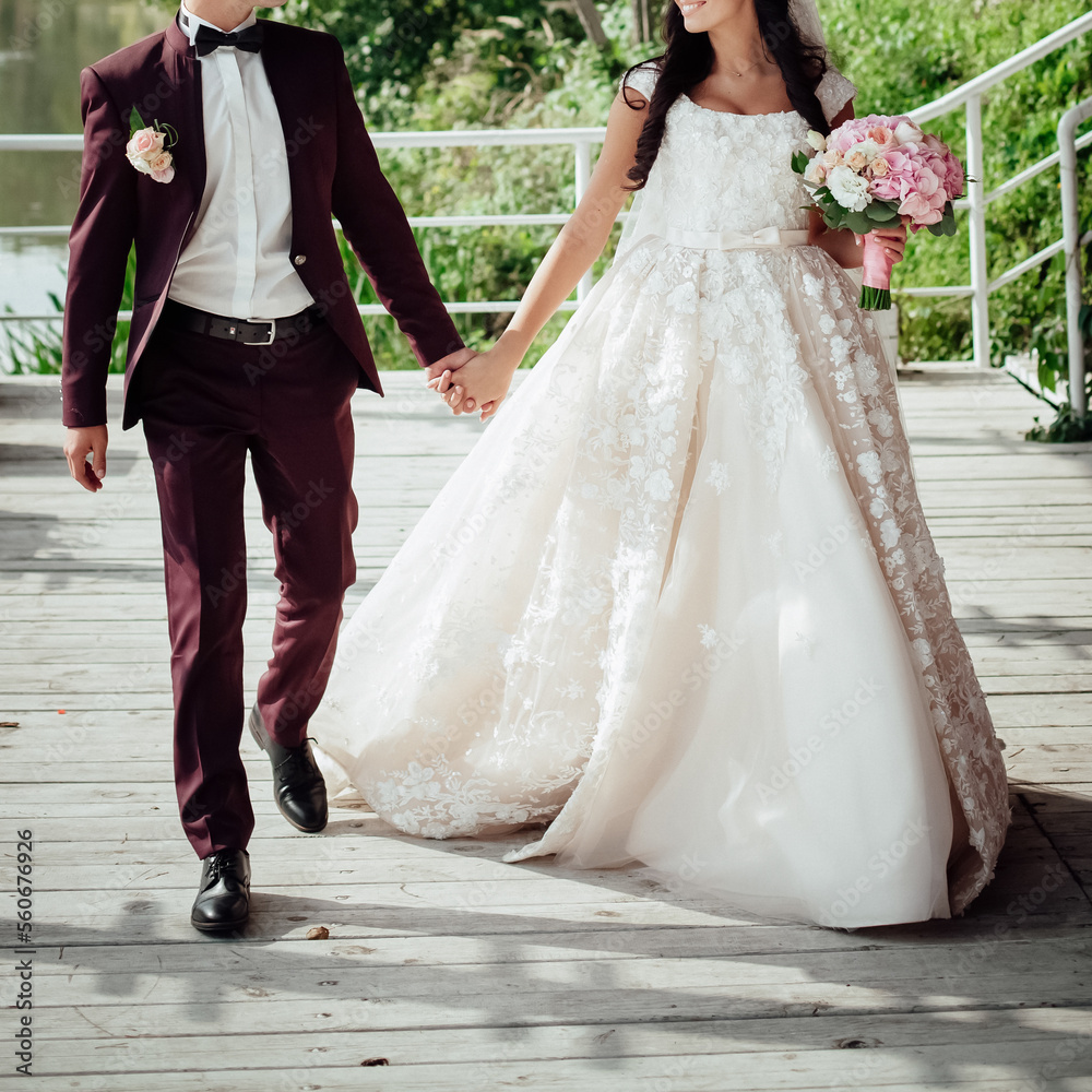 The bride and groom walk hand in hand on the pier near the river. The groom is in a crimson suit with a boutonniere, the bride is dressed in a light dress with patterns, with a bouquet in her hands. The bride and groom walk hand in hand on the pier near the river. The groom is in a crimson suit with a boutonniere, the bride is dressed in a light dress with patterns, with a bouquet in her hands.