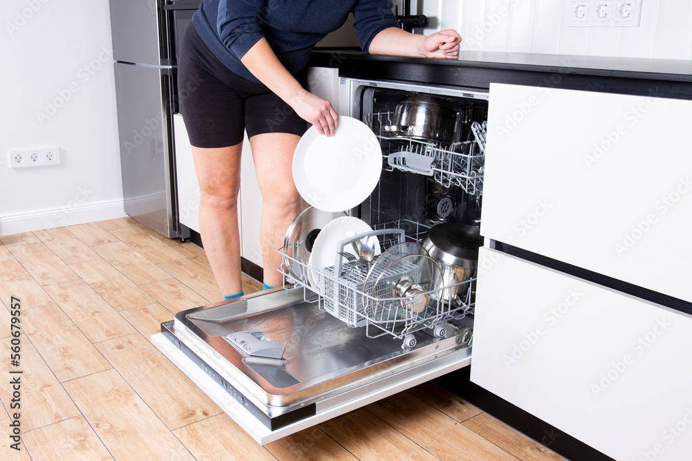 Woman is holding the clean dish with dishwasher after washing cycle, cropped image. Opened dishwasher with clean dishes.
