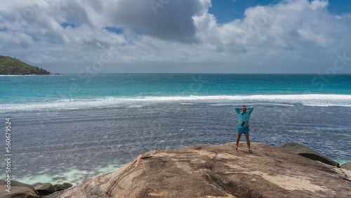 A man stands on the edge of a granite cliff, above the ocean, smiling. His arms are thrown behind his head, clothes are fluttering in the wind. Foam of waves on turquoise water. Clouds in the blue sky