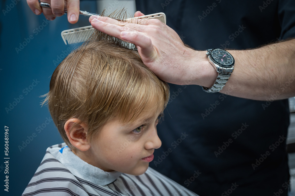 Fototapeta premium Happy cute fair-haired preschool boy getting a haircut. Children's hairdresser with scissors and comb cuts a little boy's hair in a room with a loft interior