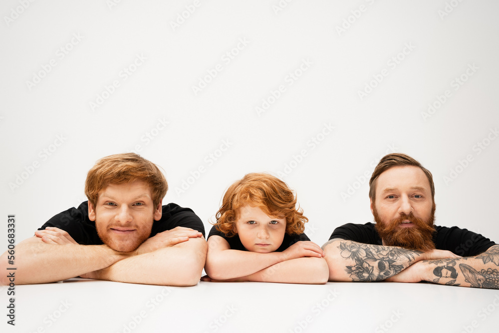 cheerful man with redhead son and bearded tattooed father lying with crossed arms on light grey background. cheerful man with redhead son and bearded tattooed father lying with crossed arms on light grey background.