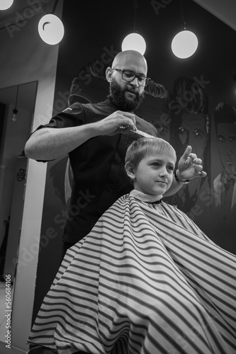 Happy cute fair-haired preschool boy getting a haircut. Children's hairdresser with scissors and comb cuts a little boy's hair in a room with a loft interior