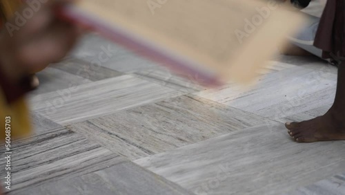 Defocus Close up of a hand holding a Sadhana buddhist prayer text. Background people walking barefoot on a white marble floor. Buddhist pilgrims doing Kora at Mahabodhi temple in Bodh Gaya, India