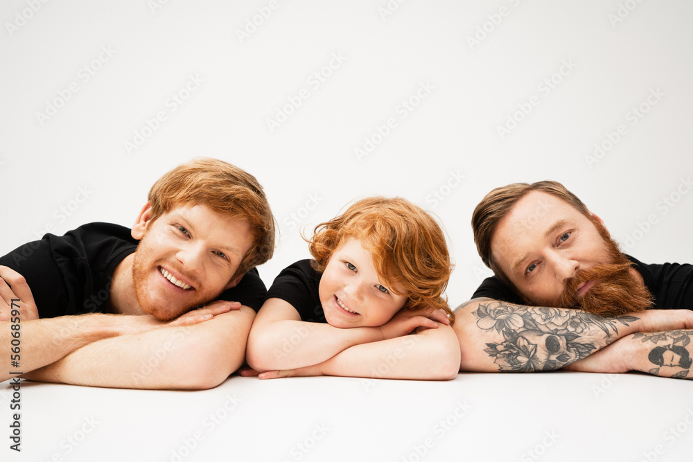 bearded tattooed man with redhead grandson and happy son smiling at camera while lying on light grey background. bearded tattooed man with redhead grandson and happy son smiling at camera while lying on light grey background.