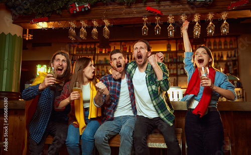 Group of young people, men and women watching match at pub. Fans emotionally cheering up favourite sport team. Celebrating winning game