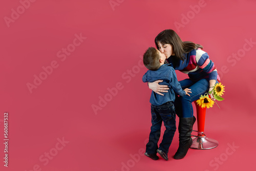 The mother kisses her son after receiving some sunflowers as a gift on a dark pink background.