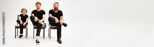 full length of bearded men and redhead boy in black clothes sitting on chairs on grey background, banner.