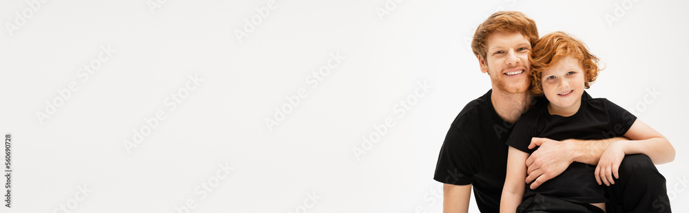 cheerful man in black t-shirt embracing redhead son and looking at camera isolated on grey, banner. cheerful man in black t-shirt embracing redhead son and looking at camera isolated on grey, banner.